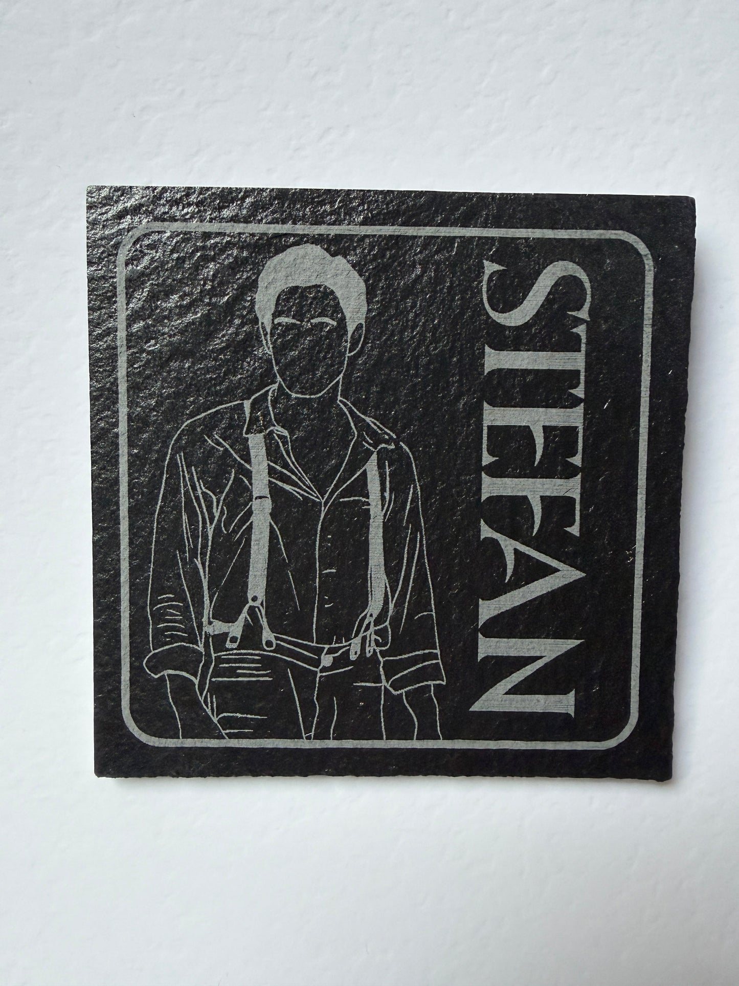 A square black slate coaster featuring a white engraved illustration of a man and the name 'STEFAN' in large white letters, displayed on a white textured background.