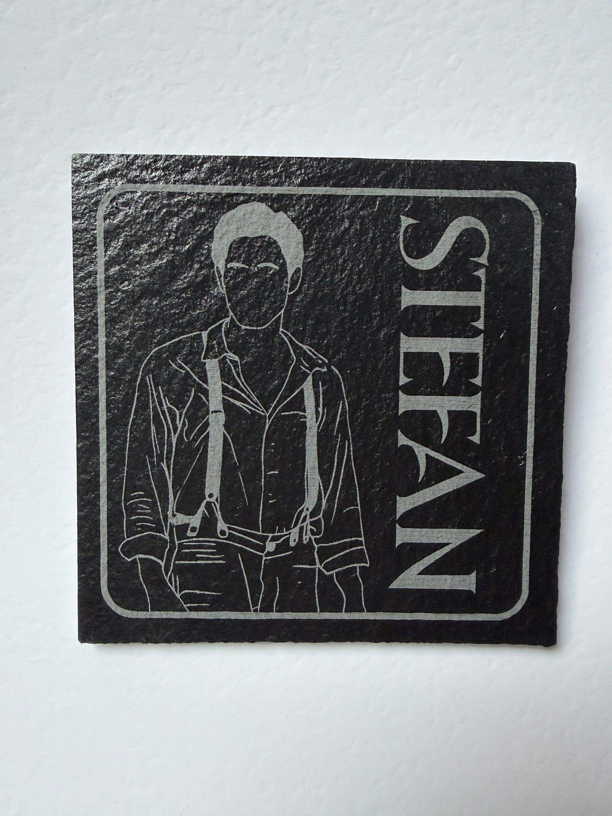 A square black slate coaster featuring a white engraved illustration of a man and the name 'STEFAN' in large white letters, displayed on a white textured background.
